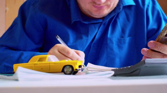 Close up of hands using calculator and holding smartphone at office desk. Car insurance agent estimating policy cost. Yellow toy car on table, shallow depth of field, bright natural light.