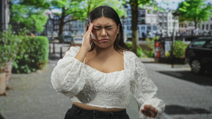 Fototapeta premium Woman pinches nose on street wearing white embroidered cropped top with hand on hip and eyes closed, near parked car and hedge; discomfort concern.
