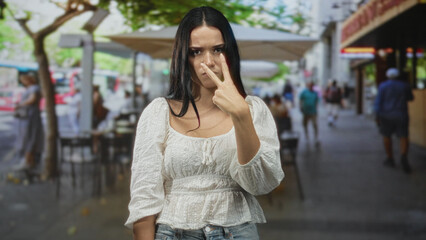 Fototapeta premium Young hispanic woman points finger directly at camera, stern expression, wearing white blouse at a sidewalk cafe on a busy city street; anger confrontation.