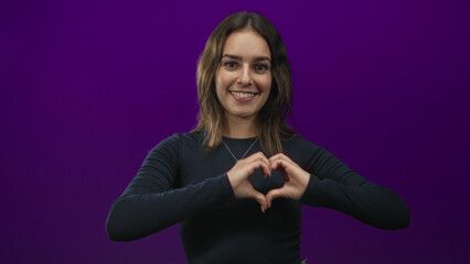 Obraz premium Young woman smiling and forming a heart with both hands in studio with deep purple backdrop; love kindness.
