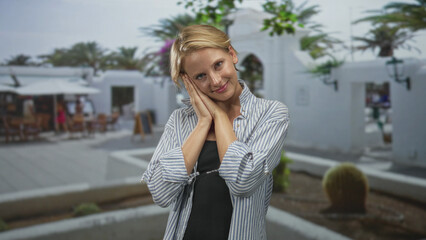 Fototapeta premium Caucasian blond woman presses hands to cheek, eyes closed in a gentle resting pose in sunlit street courtyard with cafes; serenity.