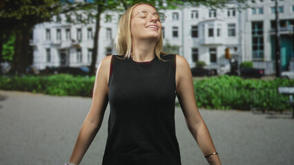 Fototapeta premium Woman tilts face upward, eyes closed and smiling, shoulders bare while standing on a leafy urban street in front of white townhouses; serenity.