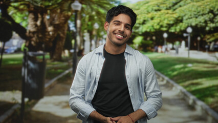 Fototapeta premium Man smiling with hands clasped at waist on a tree lined street path, wearing light shirt over black tee; warmth contentment.