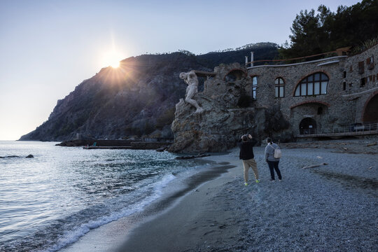 Vista di un titano scolpito che emerge da aspre scogliere a picco sulla spiaggia serena, dove le acque turchesi incontrano la riva sabbiosa; Monterosso al Mare, Liguria, Italia.