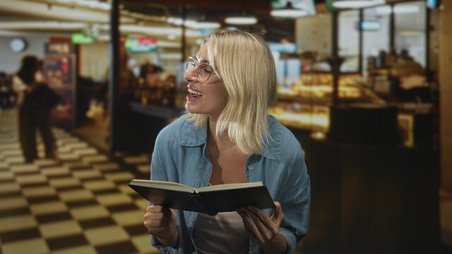 Woman reading book, smiling and holding the open hardcover while standing in a busy building cafe by a bakery counter and checkered floor; relaxed contentment.