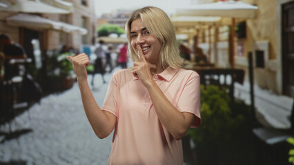Fototapeta premium Young blonde woman finger to lips for silence and thumb pointed back wearing pink polo on a cobblestone street; playful secrecy.