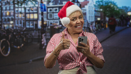 Fototapeta premium Woman in santa hat holds smartphone and gives thumbs up on a street lined with canal buildings, smiling and checking her phone; holiday joy.