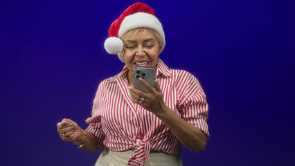 Fototapeta premium Woman senior hispanic in red striped shirt and santa hat taps smartphone with both hands and looks at screen in studio; holiday cheer.