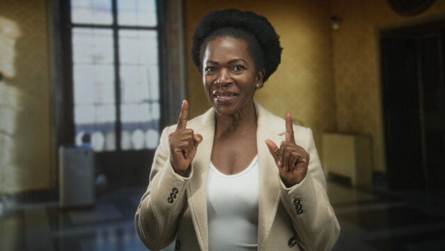 Woman points finger, hands visible, wearing beige coat and white top in a building; confidence encouragement warmth.