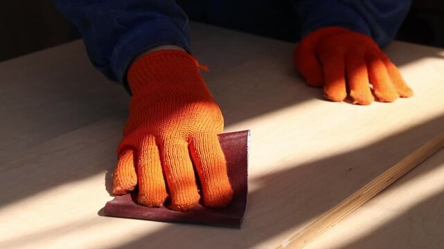 The process of manual sanding of light plywood or wooden board. A person in blue work clothes and bright orange gloves processes wood with sandpaper. The concept of carpentry, repair and woodworking