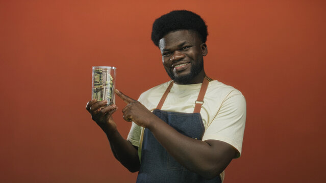 Man points finger to clear glass jar of rolled cash and dollar bills on orange studio wall; financial confidence savings.