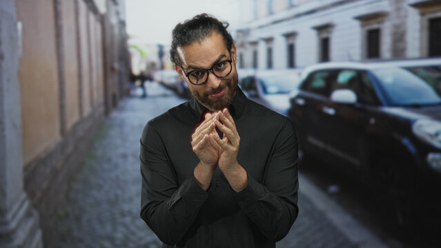 Man with glasses rubbing fingers together in black shirt on cobblestone street; scheming slyness cunning intent.