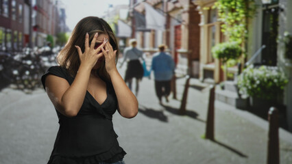 Fototapeta premium Woman covering mouth with hands on street, wearing black blouse and glasses near bollards and storefronts; shock vulnerability.