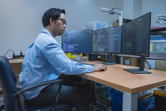 Professional Asian male software developer programming complex code across multiple computer monitors inside a modern office environment for commercial technology and IT recruitment campaigns.