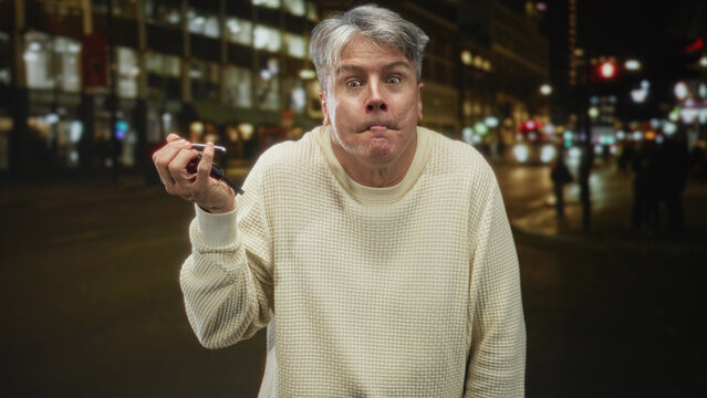 Man holding car keys in hand with pursed lips and slight shrug on a city street at night; late night confusion.