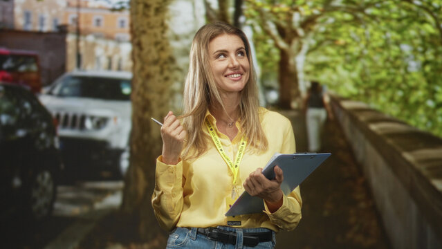 Woman holds clipboard and pen, smiles wearing visitor card lanyard and yellow shirt on street; friendly community support.