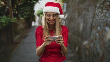 Fototapeta premium Blonde woman holding smartphone with both hands, smiling and looking down while wearing a santa hat on a narrow street; festive joy.
