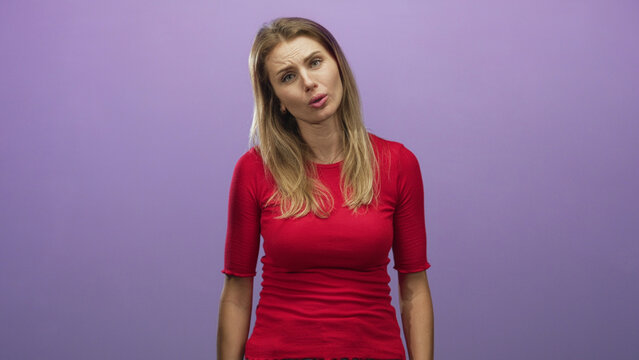 Woman presses hands together near chin, wearing red top with blonde hair visible, head tilted against purple studio backdrop; apologetic sincerity.