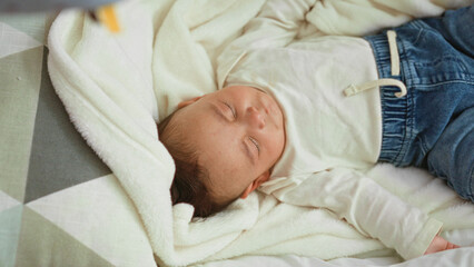 Baby boy sleeping with closed eyes and hand on soft blanket on bed in building, wearing white shirt and denim shorts  peaceful comfort. © Krakenimages.com