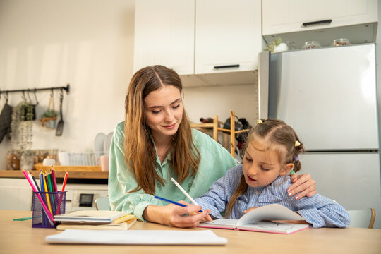 Mother helping her little daughter with homework and writing in a notebook at kitchen table. Young woman homeschooling a cute girl, explaining a lesson during supportive family study time at home.