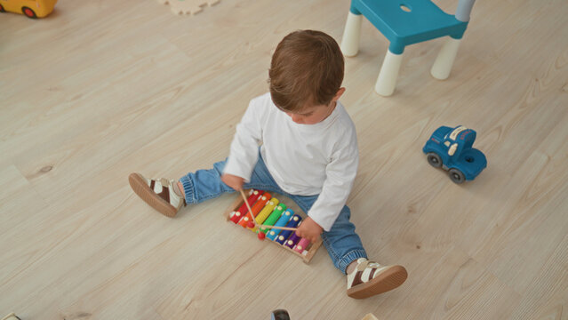 Boy sitting on wooden floor plays colorful xylophone with mallet near toy car and small chair in play area inside building; playful curiosity.