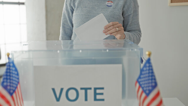 Woman inserting ballot into transparent ballot box on a table in a building polling station, flanked by small american flags; civic duty hopeful.