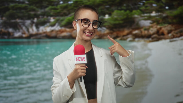 Woman holding red microphone labeled news points index finger to cheek while smiling at camera on a sandy beach cove wearing glasses white blazer black crop top; broadcast confidence.