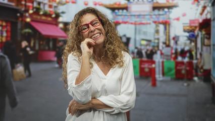Fototapeta premium Woman with hand on chin, red glasses and white blouse standing in a busy street market; thoughtful reflection.