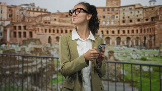 Woman holding smartphone with hands, glancing sideways in front of building wearing olive blazer and glasses; curiosity travel discovery.