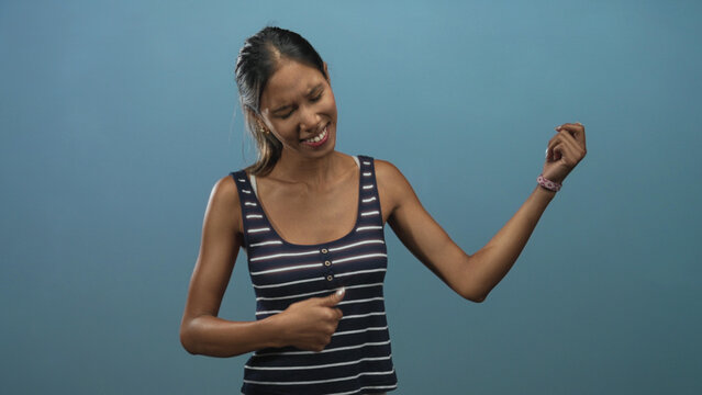 Thai woman with exposed arms miming air guitar and grimacing in studio against blue backdrop wearing striped tank top; playful energy.