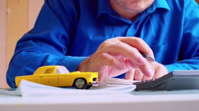 Businessman using calculator and writing notes in logbook at desk. Focus on hands, yellow toy car on table. Close up, shallow depth of field, natural light. Automotive accounting workflow.