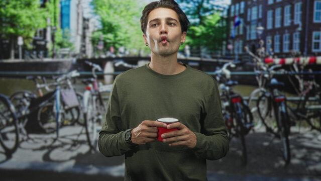 Man holding red cup purses lips on street by canal with bicycles and bridge, wearing green shirt, earring and watch for a casual moment; playful pause.