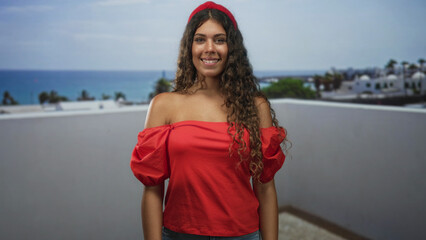 Obraz premium Teenage hispanic woman with bare shoulders in a red off shoulder top smiling and looking up on a rooftop building terrace by the sea; carefree joy.