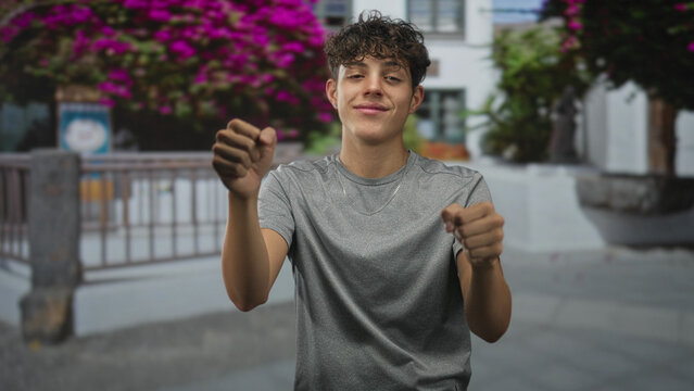 Teenage man lifts open hand to block his face while miming steering with the other hand in street near flowering bushes and railing; playful energy.
