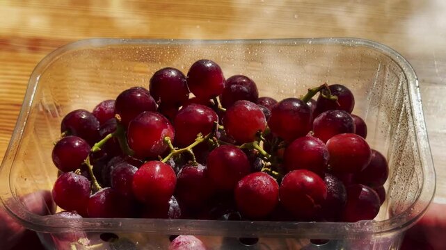 Closeup of hands pushing a punnet of fresh red grapes from a shaded area into the sunlight, on a pine counter.
