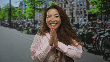 Fototapeta premium Woman with palms pressed together and smiling at camera on busy street in amsterdam wearing pink shirt and showing hands with red nails; gratitude joy.