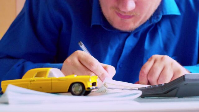 Slow zoom out on focused car dealer hands computing and writing in ledger. Yellow toy car on table, close up, selective focus, shallow depth of field, natural daylight. 