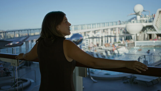 Woman rests arm on deck railing and smiles while gazing out at the ship pool and promenade; serenity.