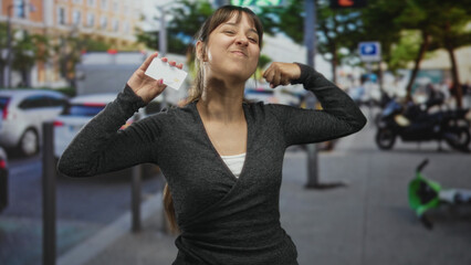 Fototapeta premium Young hispanic woman holds creditcard up and flexes arm on city street while smiling with eyes closed; confidence success.