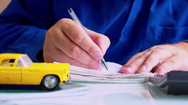 Slow zoom out on focused businessman writing results in ledger. Focus on hands, yellow toy car on desk. Shallow depth of field, natural daylight. Automotive accountant workflow.