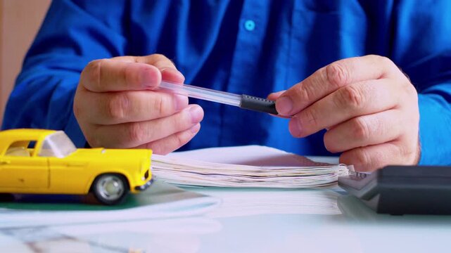 Businessman twirling pen at office desk close up. Focus on hands. Yellow toy car on table. Shallow depth of field, natural daylight. Bored automotive entrepreneur procrastinating.