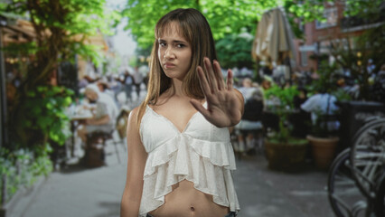 Naklejka premium Woman with hand raised palm out on street cafe terrace showing bare midriff and scowl; boundary defiance.