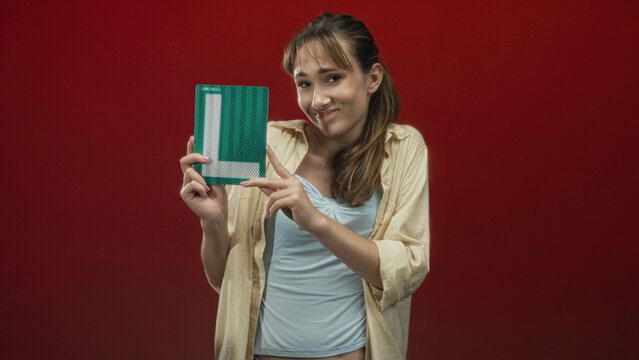 Woman holds green learner sign and points finger to it against a red studio backdrop; sheepish pride.