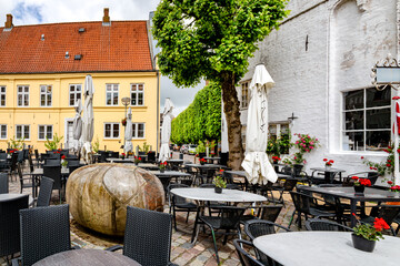 Obraz premium Cafe terrace in the heart of Tønder with cobblestones, historic buildings, large trees, umbrellas and a cozy old town atmosphere beneath a blue sky with clouds.