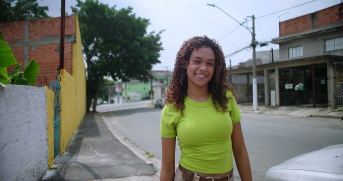 Hispanic teenager of African descent smiling warmly while walking on a quiet street in a South American urban city, natural curly hair, casual style, vibrant and cheerful