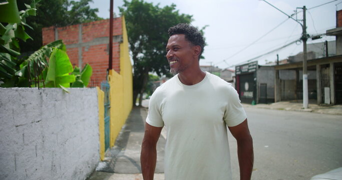 Man of African descent smiling confidently on a quiet street in a South American urban city, casual and relaxed, surrounded by greenery and colorful buildings