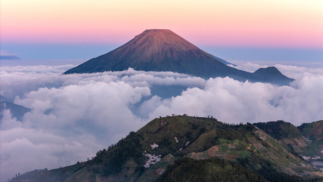 Aerial view of Mount Sindoro rising above a dense sea of clouds with soft pink and orange light at sunrise in Indonesia.