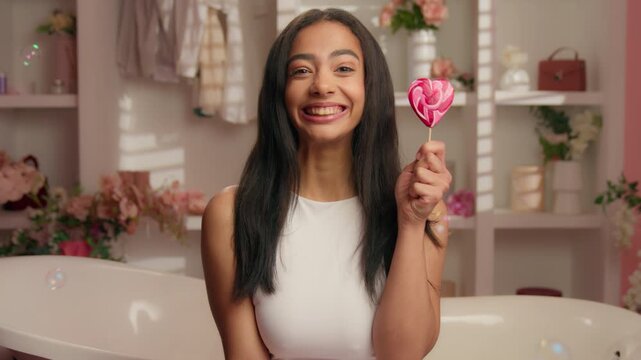Cheerful American girl holding candy heart shape smiling in pink bathroom flying soap bubbles Hispanic young woman Latina ethnic female smile toothy with white teeth dental stomatology medicine health
