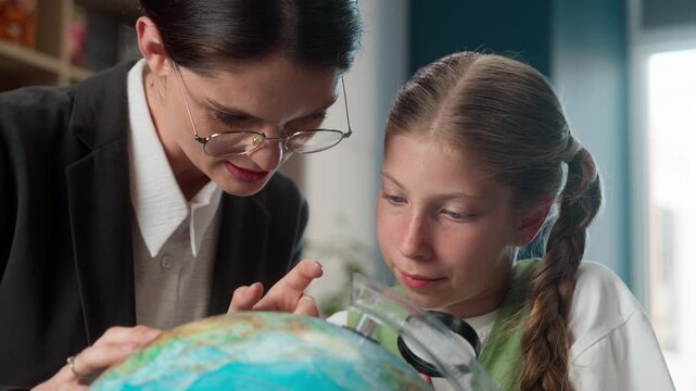 Caucasian teacher tutor woman teaching little school girl child kid daughter pupil in class looking at globe together talking studying learning geographic study learn look at earth world map education