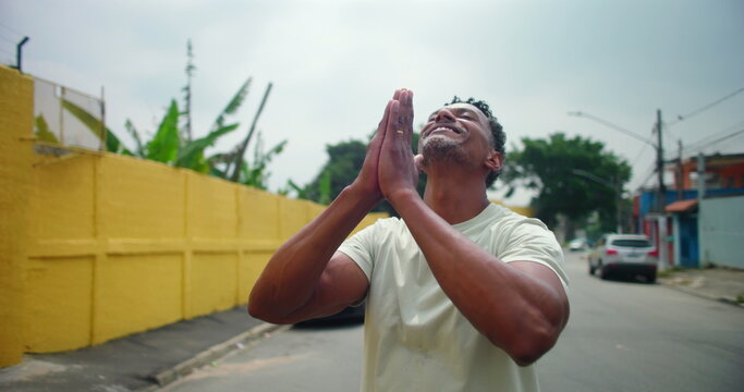 Man of African descent in an urban street, hands clasped in prayer, looking upward with a peaceful expression, symbolizing spirituality, hope, and devotion